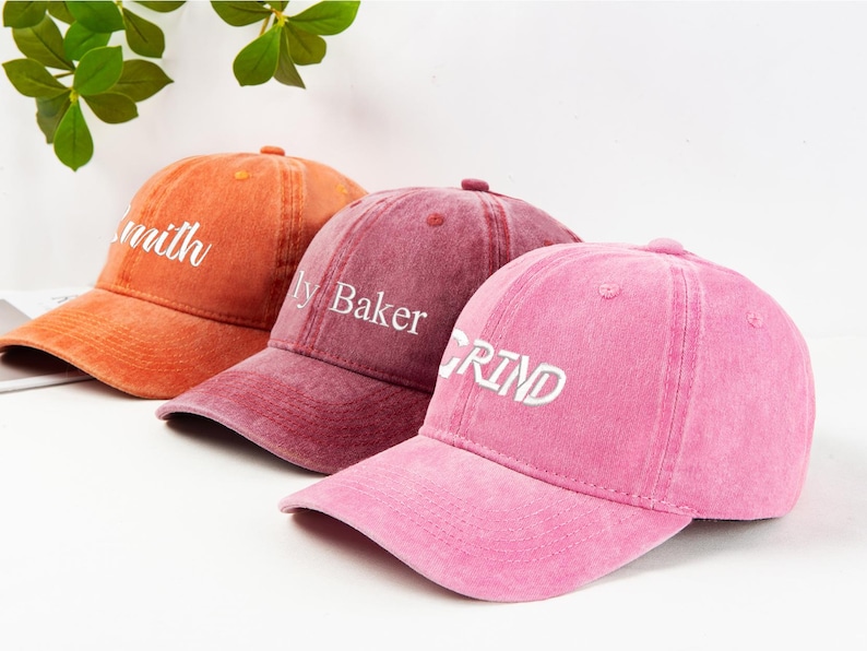 May include: Three baseball caps in shades of orange, burgundy, and pink. Each cap has a curved brim and a top button. The orange cap reads "Smith", the burgundy cap reads "ly Baker", and the pink cap reads "GRIND" in white lettering.