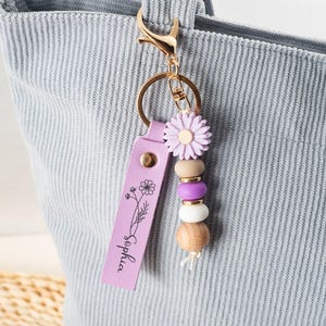 May include: A lavender leather keychain with the name "Sophia" and a floral design. The keychain features a gold clasp, a daisy-shaped charm, wooden and purple beads, and a tassel.