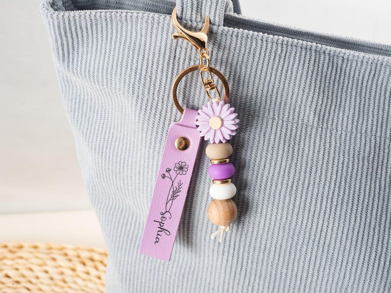 May include: A lavender leather keychain with the name "Sophia" and a floral design. The keychain features a gold clasp, a daisy-shaped charm, wooden and purple beads, and a tassel.