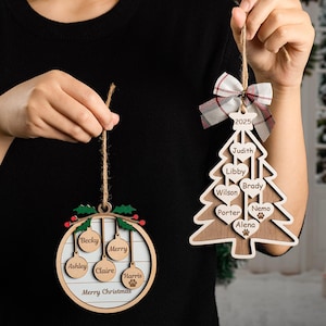 May include: Two wooden Christmas decorations are held up. One is a circle with names and "Merry Christmas". The other is a tree with names and "2025". Both are made of light wood and have twine for hanging.