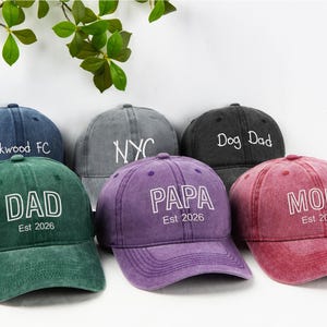 May include: A collection of six baseball caps in various colors, including green, purple, and red. Each cap features white text, with phrases like "DAD", "MOM", and "PAPA", and the year "Est 2026". The caps are arranged on a white surface.