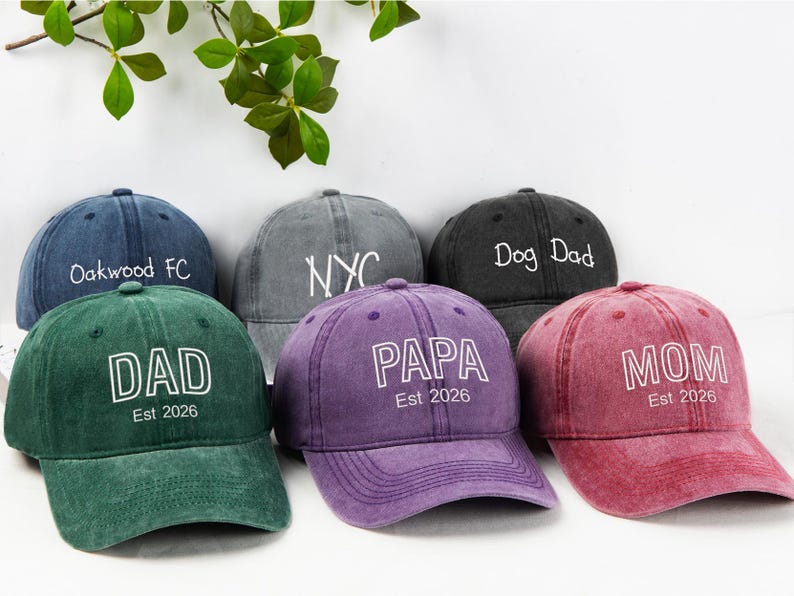 May include: A collection of six baseball caps in various colors, including green, purple, and red. Each cap features white text, with phrases like "DAD", "MOM", and "PAPA", and the year "Est 2026". The caps are arranged on a white surface.