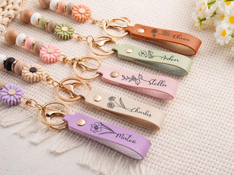 May include: Close-up of several personalized leather keychains in various colors, including orange, green, pink, beige, and purple. Each keychain features a name and floral design. The keychains are attached to beaded accessories with flower accents.
