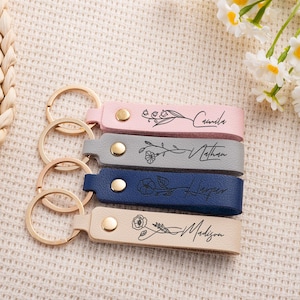 May include: Four personalized leather keychains in pink, gray, navy blue, and beige. Each keychain features a gold-colored ring and a name with floral accents. The names are Camila, Nathan, Harper, and Madison.