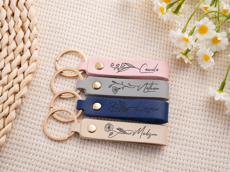 May include: Four personalized leather keychains in pink, gray, navy blue, and beige. Each keychain features a gold-colored ring and a name with floral accents. The names are Camila, Nathan, Harper, and Madison.