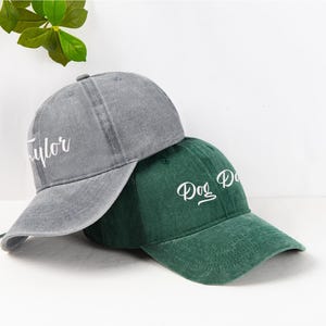 May include: Two baseball caps, one gray and one green, are displayed. The gray cap has the name "Taylor" embroidered in white, while the green cap says "Dog Dad". Both caps have a curved brim and a classic baseball cap design.