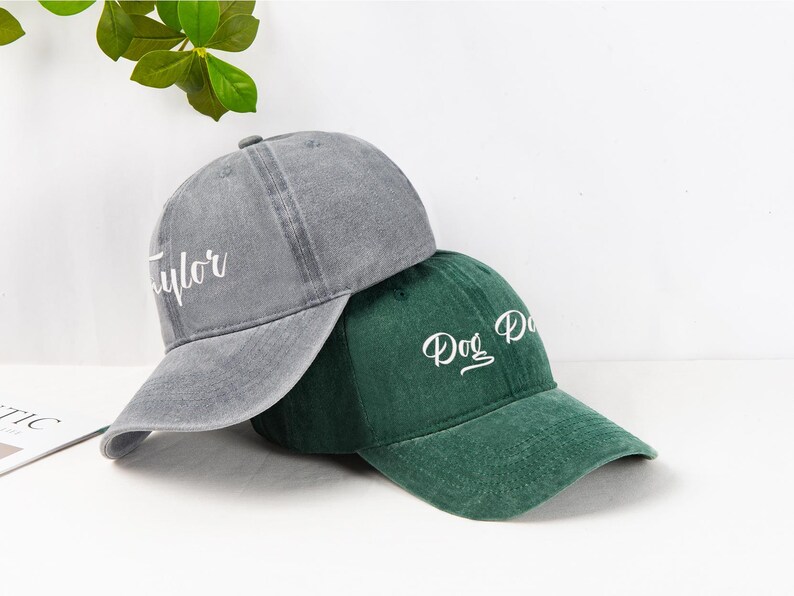 May include: Two baseball caps, one gray and one green, are displayed. The gray cap has the name "Taylor" embroidered in white, while the green cap says "Dog Dad". Both caps have a curved brim and a classic baseball cap design.