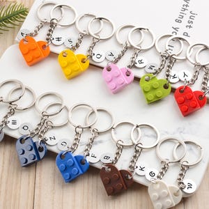 Pode incluir: Uma coleção de chaveiros coloridos em forma de coração. Cada chaveiro apresenta um design de coração de duas peças em várias cores, incluindo laranja, amarelo, rosa, verde, vermelho, cinza, azul e marrom. Cada chaveiro tem um anel e uma corrente prateados, com um pequeno pingente de letra.