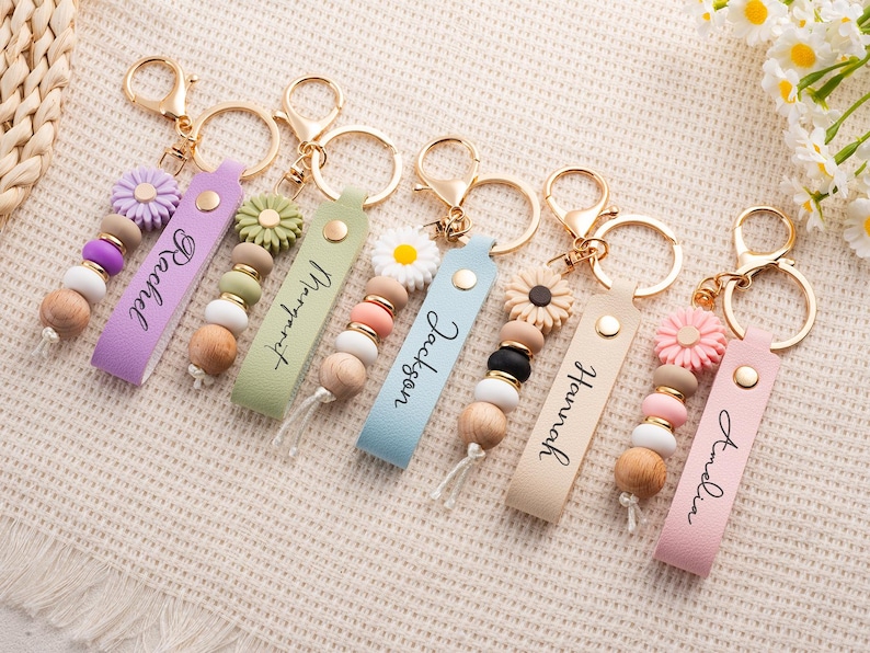 May include: A collection of personalized keychains with gold-colored hardware. Each keychain features a leather strap with a name, wooden beads, and a decorative flower. The keychains are in various colors, including purple, green, blue, beige, and pink.