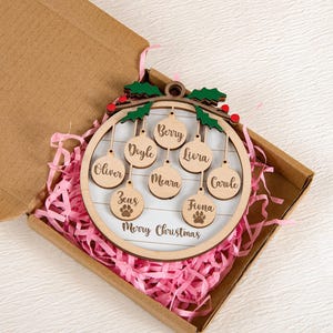 May include: A wooden Christmas ornament with a circular frame and hanging name tags. The ornament includes names and a paw print, with the words "Merry Christmas" at the bottom. It is presented in a cardboard box with pink paper.