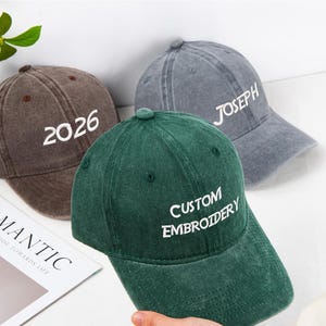 May include: Three baseball caps in brown, gray, and green. The brown cap has "2026" embroidered in white. The gray cap has "JOSEPH" embroidered in white. The green cap has "CUSTOM EMBROIDERY" embroidered in white. All caps have a curved brim.