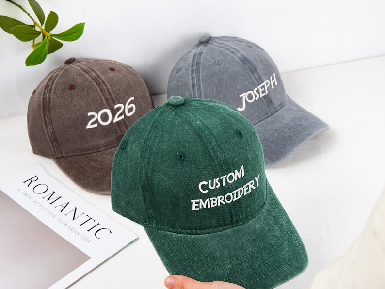 May include: Three baseball caps in brown, gray, and green. The brown cap has "2026" embroidered in white. The gray cap has "JOSEPH" embroidered in white. The green cap has "CUSTOM EMBROIDERY" embroidered in white. All caps have a curved brim.