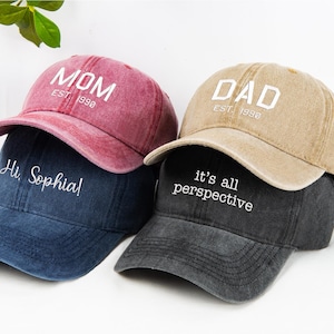 May include: A stack of four baseball caps in shades of red, blue, tan, and gray. The red cap says "MOM EST 1998", the blue cap says "Hi Sophia!", the tan cap says "DAD EST 1990", and the gray cap says "it's all perspective".