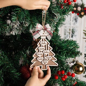 May include: A wooden Christmas tree ornament with names engraved on heart-shaped cutouts. The ornament is held in front of a Christmas tree. A plaid bow and the year 2025 are at the top. The names include Stella, Isabel, George, Carter, Kathie, Daniel, and Elanie.