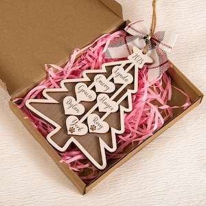 May include: A wooden Christmas tree ornament with a light brown base and white outline, personalised with names on heart-shaped accents. The ornament is in a cardboard box with pink paper filler, and a plaid bow.
