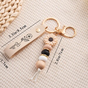 May include: A beige leather keychain with gold-colored hardware. The keychain features a floral design and the name "Bella". It has a sunflower charm and a beaded accent with wooden, black, and white beads. The leather strap is 3.3 inches long.