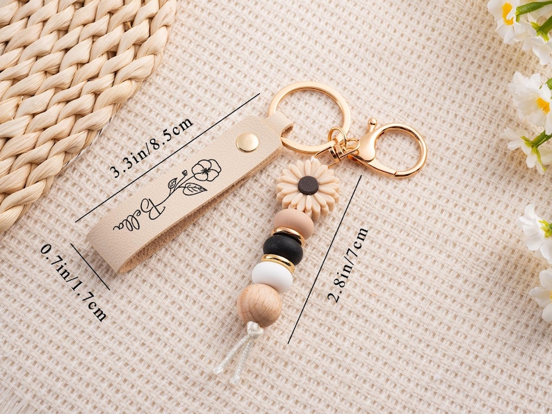 May include: A beige leather keychain with gold-colored hardware. The keychain features a floral design and the name "Bella". It has a sunflower charm and a beaded accent with wooden, black, and white beads. The leather strap is 3.3 inches long.