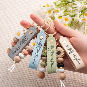 May include: A hand holds five personalized keychains with leather straps in gray, blue, green, and beige. Each keychain features a floral design and a name. The keychains have wooden beads, daisy accents, and gold-tone hardware.