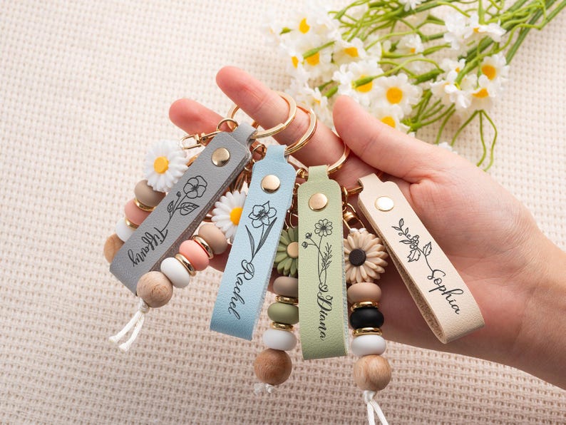 May include: A hand holds five personalized keychains with leather straps in gray, blue, green, and beige. Each keychain features a floral design and a name. The keychains have wooden beads, daisy accents, and gold-tone hardware.