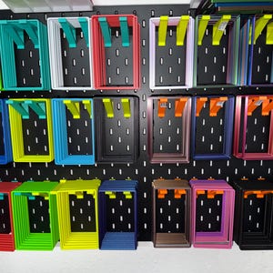 Könnte beinhalten: Eine Präsentation von farbenfrohen, rechteckigen Aufbewahrungsbehältern mit abgewinkelten Halterungen, die auf einer schwarzen Lochwand montiert sind. Die Behälter sind in verschiedenen Farben erhältlich, darunter Grün, Blau, Rot, Gelb und Rosa. Die Lochwand hat gleichmäßig verteilte Löcher.