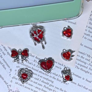 May include: A collection of six silver charm pendants with red glass hearts. The charms feature different designs, including a broken heart, a heart with barbed wire, a heart with flames, a heart with a bow, and a heart with a floral design.