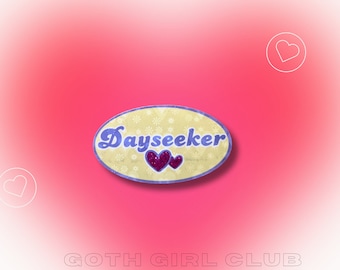 Pode incluir: Um adesivo oval amarelo com o texto "Dayseeker" em letras roxas e dois corações rosa com glitter. O adesivo tem um fundo branco com um padrão floral. O adesivo está sobre um fundo rosa com o texto "GOTH GIRL CLUB" em letras brancas.