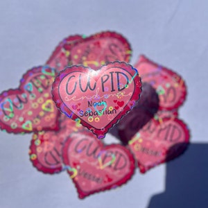 May include: A stack of pink heart-shaped stickers with a holographic finish. The top sticker says "CUPID sends love Noah Sebastian".