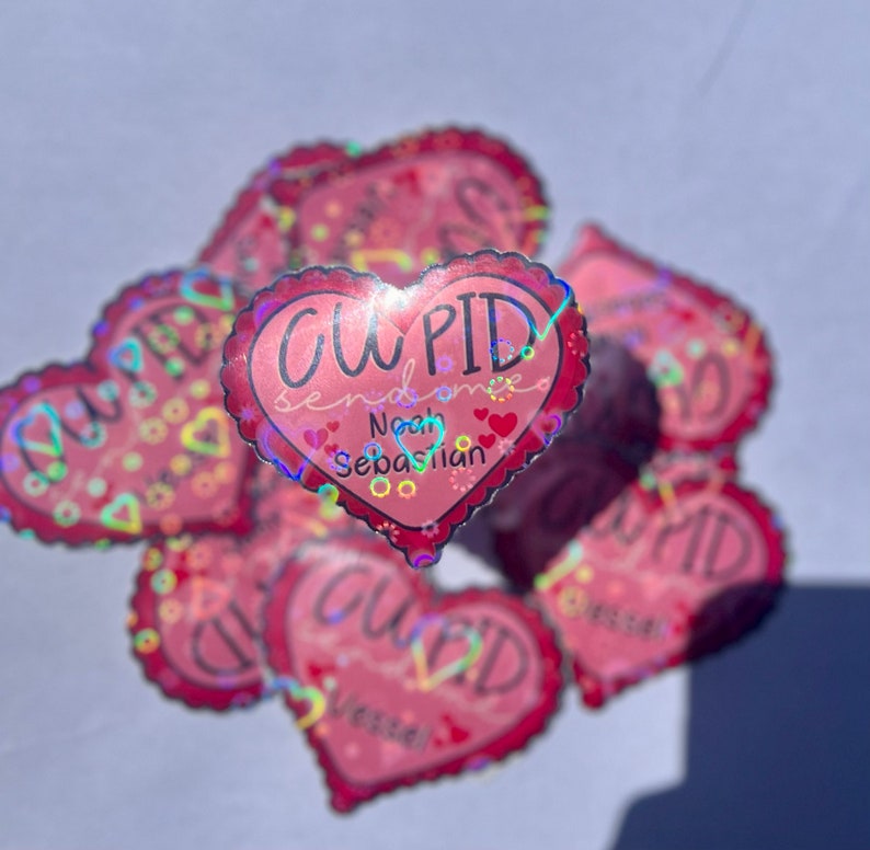 May include: A stack of pink heart-shaped stickers with a holographic finish. The top sticker says "CUPID sends love Noah Sebastian".