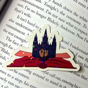 May include: A sticker of a book with a gothic cathedral on the cover. The book is open and the cathedral is glowing with a magical effect. The sticker is on a page of a book with text that reads "Running my hands down the white deep breath. Tonight's the ceremony. To sure, I'm no longer Blakely Anderson. Now I'm Ryat's wife. It isn't hard to sa also understand commitment. The fact th ough for "Blake, y Yeah," m  Ayat call out, entering his room.  as he enters. mes to a stop; his emerald eyes drop owly run up over the fitted materia a slit up my right leg. wear because. y cho"