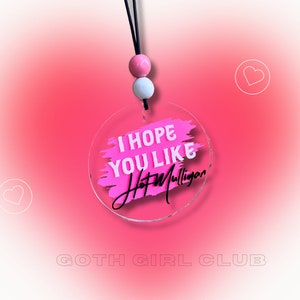 May include: A pink and white acrylic charm with a black string and beads. The charm has a pink brushstroke design with the text "I hope you like Hot Mulligan".