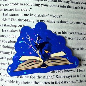 May include: A blue and silver glitter book bookmark with a black silhouette of a dragon and a tree with stars. The bookmark is on top of an open book with the text "already knows." "I lock" "Guess we're done for the night," Kaori says as a line only visible by their silhouettes in the darkness. "The s" "You two should return to your dragons."