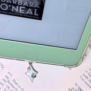 May include: A close-up of a green Kindle e-reader with a silver star and moon bookmark charm. The screen displays the title "BARBARA O'NEAL" and the text "everything about her life."