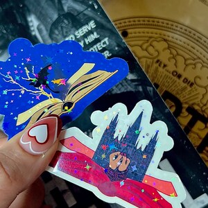 May include: Two holographic stickers with glitter. The first sticker is blue with a black silhouette of a bat flying over an open book. The second sticker is white with a black silhouette of a building with a red cape draped over it. Both stickers have a starry background.
