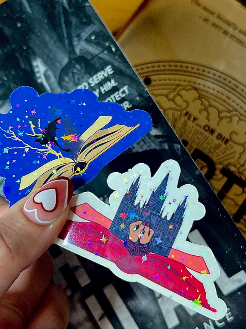 May include: Two holographic stickers with glitter. The first sticker is blue with a black silhouette of a bat flying over an open book. The second sticker is white with a black silhouette of a building with a red cape draped over it. Both stickers have a starry background.