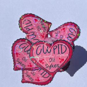 May include: A stack of pink heart-shaped stickers with a holographic finish. The stickers say "Cupid find me" and "Oli Sykes".