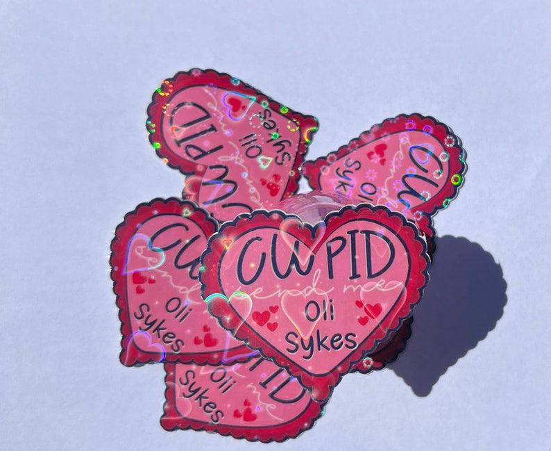 May include: A stack of pink heart-shaped stickers with a holographic finish. The stickers say "Cupid find me" and "Oli Sykes".