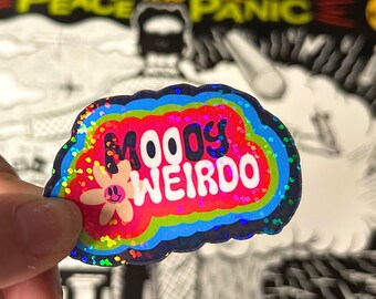 Puede incluir: Una pegatina holográfica con un borde arcoíris y un fondo rojo. La pegatina dice "MOODY WEIRDO" en letras blancas con una flor sonriente en la esquina.