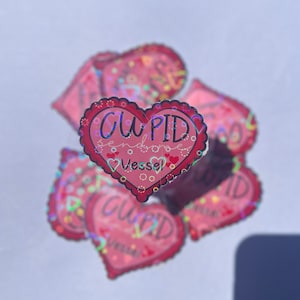 May include: A stack of pink heart-shaped stickers with a holographic finish. The sticker in the front reads "CUPID sends some Vessel".