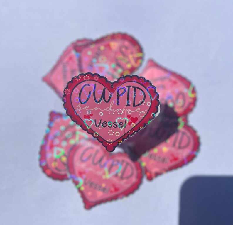 May include: A stack of pink heart-shaped stickers with a holographic finish. The sticker in the front reads "CUPID sends some Vessel".
