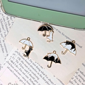 Peut inclure: Trois breloques dorées représentant des chats noirs et blancs tenant des parapluies noirs et blancs. Les breloques sont sur un fond blanc avec du texte.