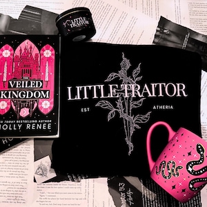 Puede incluir: Una camiseta negra con el texto "Little Traitor" y un gráfico de una rosa con espinas. El texto "Est" y "Atheria" también están en la camiseta. Una taza rosa con un diseño de serpiente y estrellas está en primer plano. Un libro titulado "The Veiled Kingdom" está en el fondo.