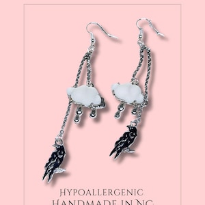 May include: Silver-toned dangle earrings with a cloud and raven design. Each earring features a white cloud with silver rain drops, a black raven, and the text "A Rainy Day". The earrings are labeled "Hypoallergenic Handmade in NC".
