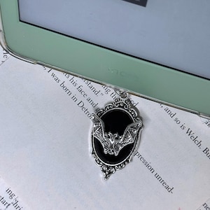 Könnte beinhalten: Silbernes Lesezeichen mit einem schwarzen Fledermaus-Design in einem ovalen Rahmen. Das Lesezeichen liegt auf einer Seite eines Buches mit Text.
