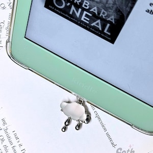 May include: A close-up of a mint green Kindle e-reader with a silver cloud-shaped bookmark with raindrops hanging from it. The book "Barbara O'Neal" is visible on the screen.