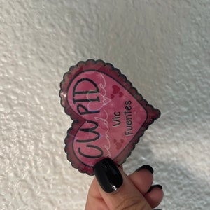 May include: A pink heart-shaped sticker with the word "CUPID" written in black and the text "send love" and "Vic Fuentes" written in white.