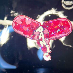 May include: A pink and red glitter sticker with the word "PANIC" in white letters. The sticker is shaped like a heart and has a glittery finish.