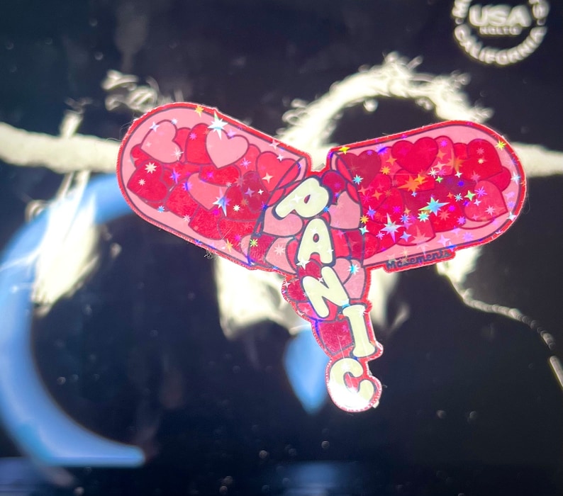 May include: A pink and red glitter sticker with the word "PANIC" in white letters. The sticker is shaped like a heart and has a glittery finish.