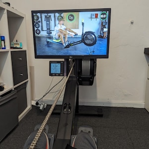 Peut inclure: Un rameur noir avec les pieds d'une personne sur les repose-pieds. La machine est équipée d'un écran numérique affichant le temps, la distance et d'autres données d'entraînement. Un écran de télévision montre une personne en train de ramer sur un rameur.