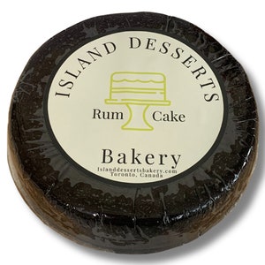 May include: A round, dark brown cake with a white label that says "Island Desserts" in a black circle with a yellow cake icon and the words "Rum Cake" below. The label also says "Bakery" with the website "islanddessertsbakery.com" and "Toronto, Canada" below.
