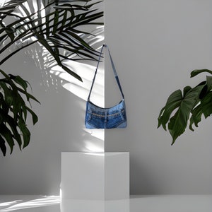 May include: A blue denim shoulder bag with a patchwork design hangs on a white wall. The bag is made from recycled denim and has a unique, handmade look.